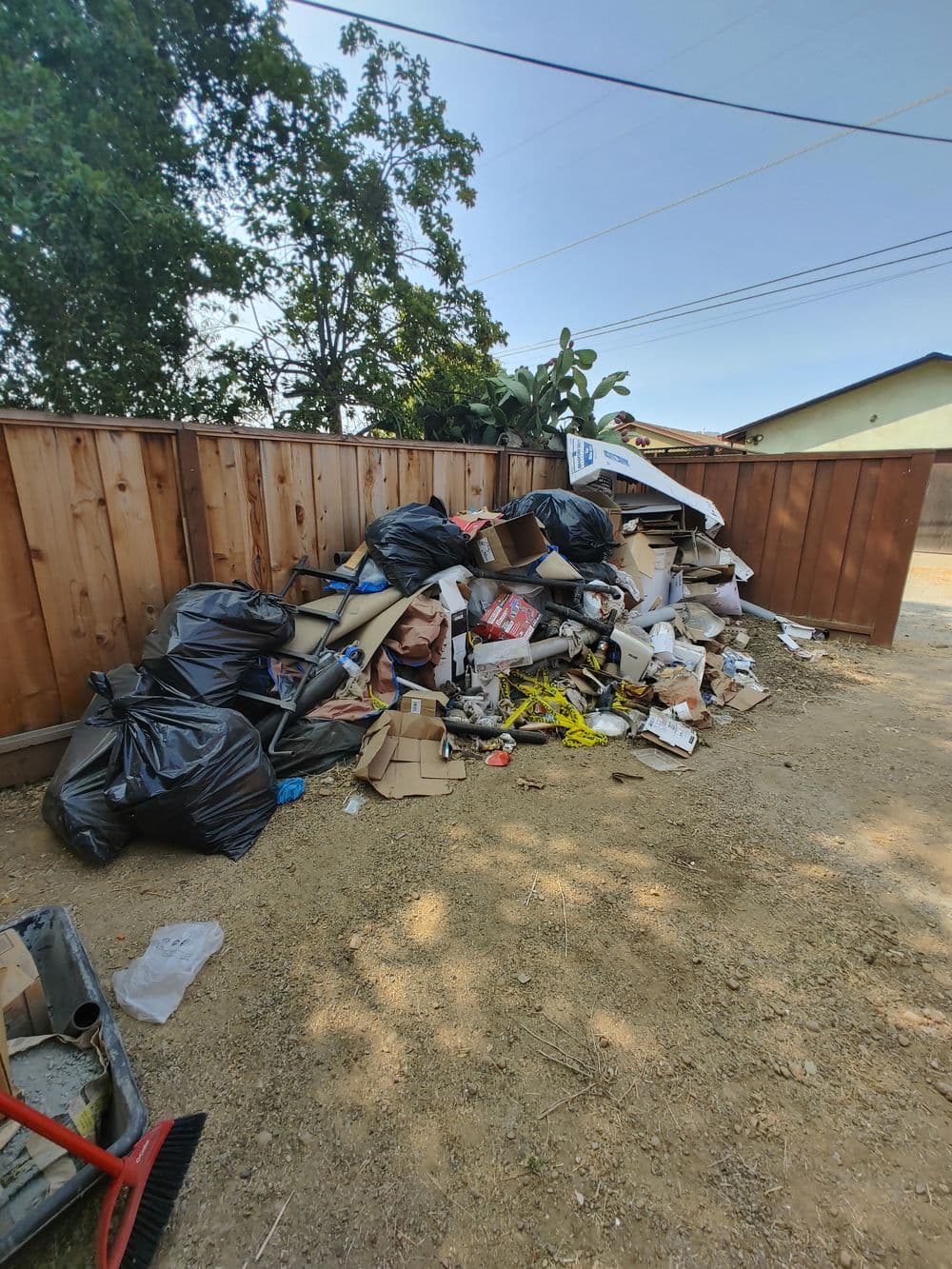 Piled-up garbage bags and debris behind a wooden fence in a cluttered outdoor area.
